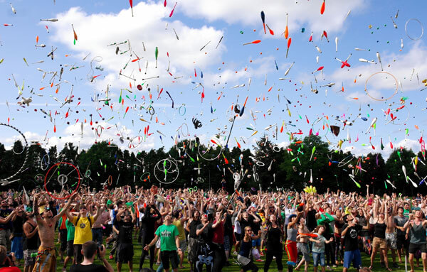 eine große Menschenmenge jongliert gleichzeitig mit Kegeln auf einer weitläufige Rasenfläche an einem Sommertag im Hintergrund dunkle Bäume und blauer Himmel mit Wölkchen