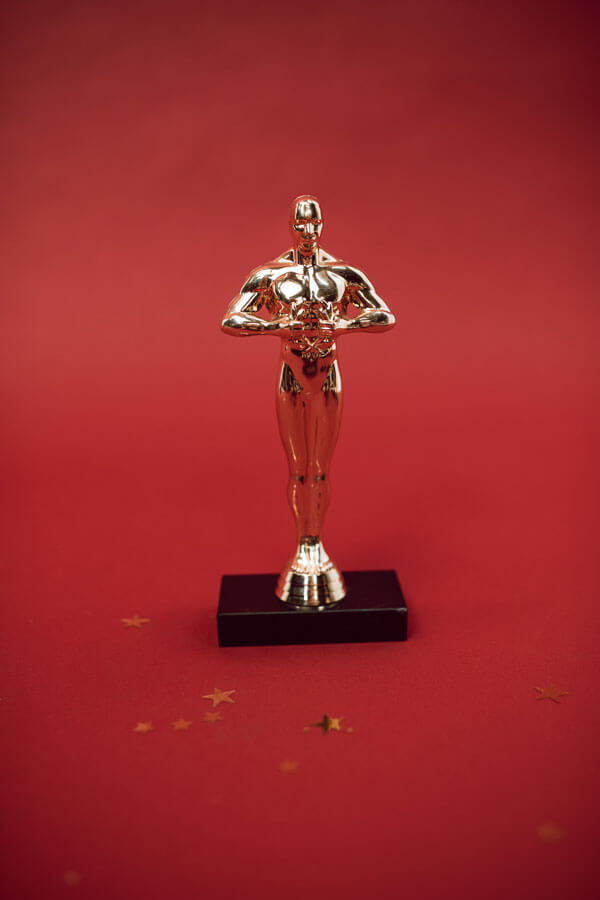 Auf rotem Unter- und Hintergrund mit goldenen Sternchen verstreut steht ein goldener Award als männliche Statue auf braunem Holzsockel