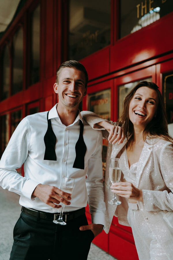 Sehr festlich gekleidetes Paar lacht und feiert mit gefüllten Sektgläsern in den Händen vor einem roten Fensterhintergrund indoor, Gäste beim Firmenjubiläum in Hamburg