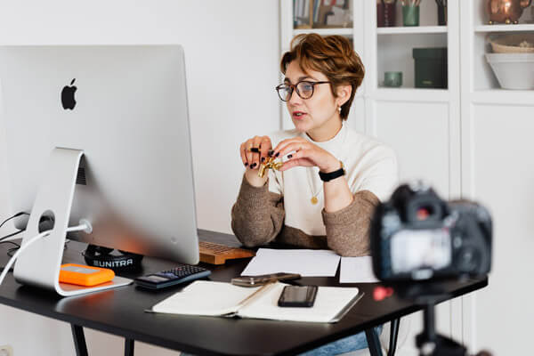 Ein Frau filmt sitzt vor einem großen Flatscreen und filmt sich mit einer Kamera unscharf im Vordergrund. Sie erklärt und schult im Homeoffice