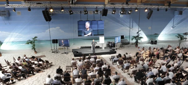 Vortrag Tagung in Halle mit Sandboden und Strand Anmutung Wind mit Südsee Motiv am Meer und Speaker auf Bühne vor Großleinwand in Szene gesetzt von Eventagentur Hamburg